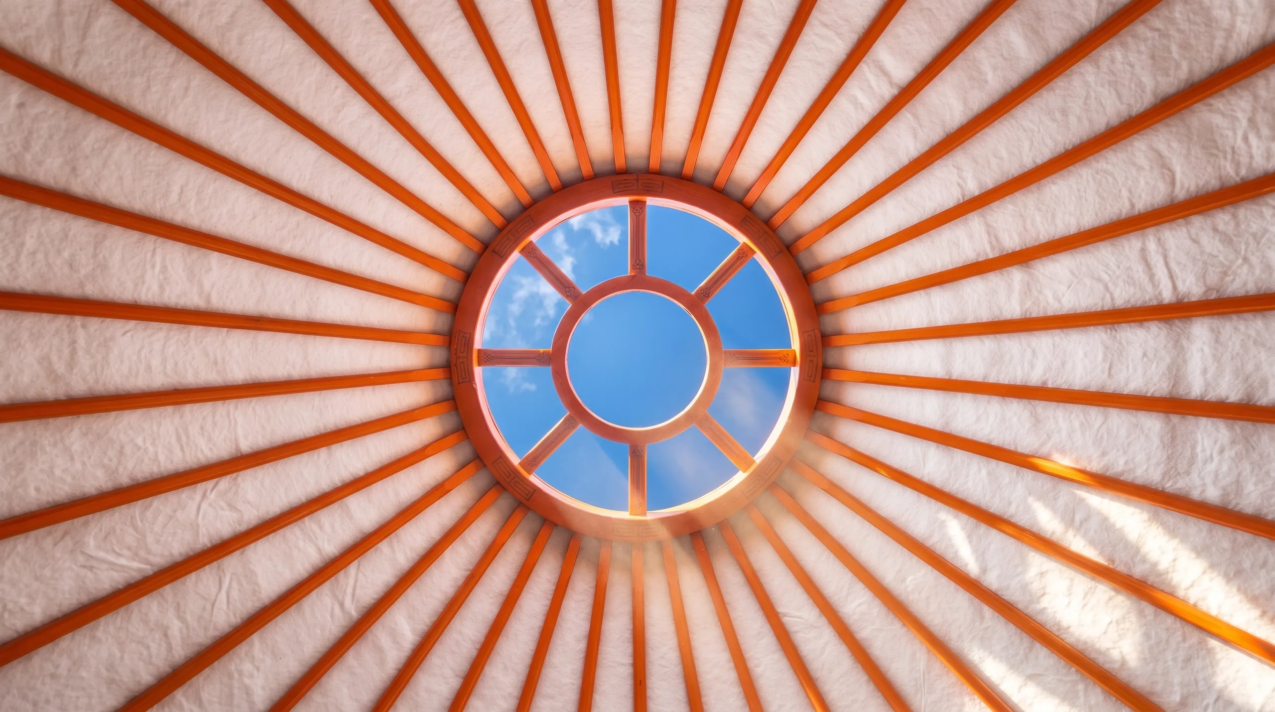 The toono crown from below, orange rafters radiating on white felt, blue sky in the opening